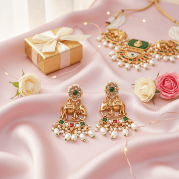 Gold earrings with green and red stones on a pink fabric background with flowers and a gift box.
